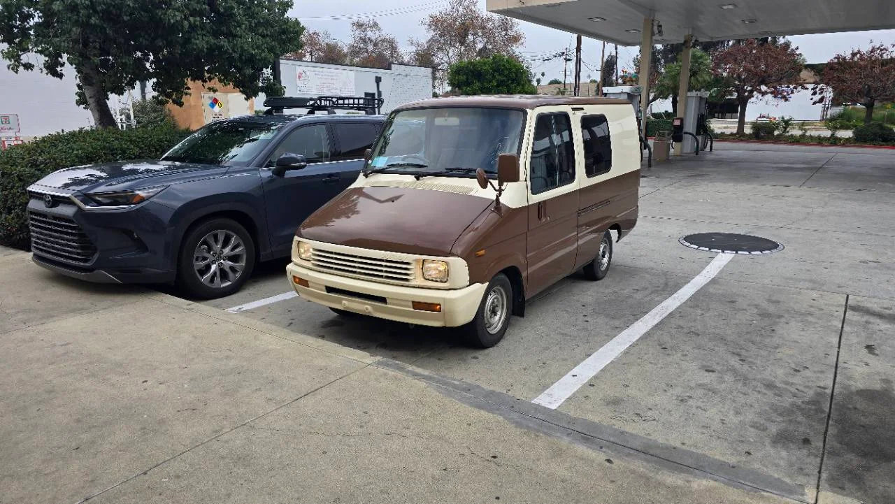 Deliboy, the cream-and-brown Toyota van, photographed from the front at a fuel stop before his store lettering